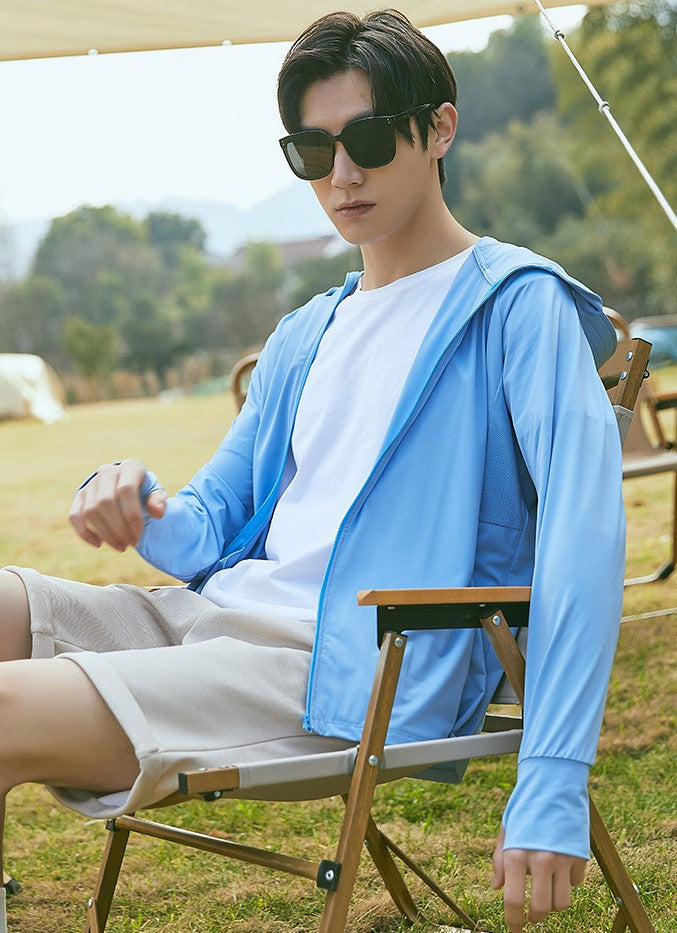 Man sitting outdoors in a chair, wearing a blue sun-protection jacket with hood and sunglasses.